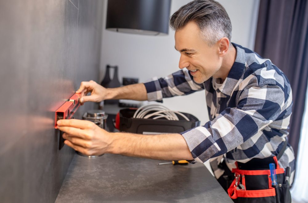 Good result. Smiling middle-aged man in plaid shirt looking approvingly and touching measuring instrument against dark smooth wall near socket in kitchen